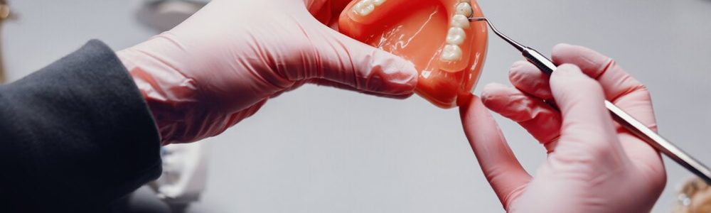 Dos manos enguantadas con guantes de latex blancos sostienen una prótesis dental casi completa sobre un paladar artificial y realiza una operación sobre un premolar